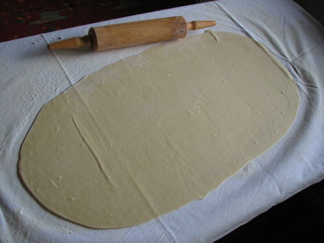<p>Tuch über den Tisch legen, mit Mehl bestäuben, Teig noch einmal kurz durchkneten, zu einem Rechteck auswalken, so weit es geht.</p>